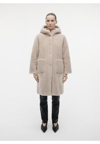 Cappotto in pelliccia sintetica beige con cappuccio, caratterizzato da una linea dritta, due tasche anteriori e una texture morbida. Abbinato a pantaloni neri e scarpe con punta.