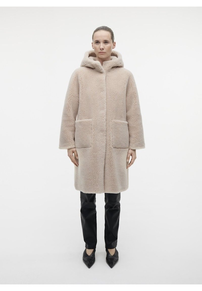 Cappotto in pelliccia sintetica beige con cappuccio, caratterizzato da una linea dritta, due tasche anteriori e una texture morbida. Abbinato a pantaloni neri e scarpe con punta.