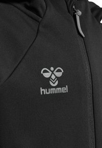 Svart sportjacka med grå dragkedja, med en framträdande grå logotyp. Slät textur med hög krage och figursydd design.