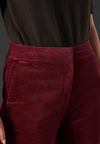 Bordowe spodnie korduroy o gładkiej fakturze, z bocznymi kieszeniami i dopasowanym pasem, zestawione z ciemną górą na zdjęciu.