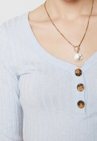 Ljusblå ribbad stickad topp med V-ringning, tre stora mörka knappar och ett guldhänge med en rund pendel.