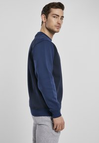 Marineblå genser med rund hals, ribbestrikkede mansjetter og kant. Har en jevn tekstur og tettsittende silhuett. Brukt sammen med grå joggebukser.