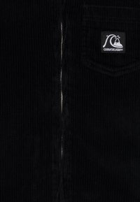 Primer plano de una tela de pana negra con una cremallera vertical y un pequeño parche del logo de Quiksilver en un bolsillo.