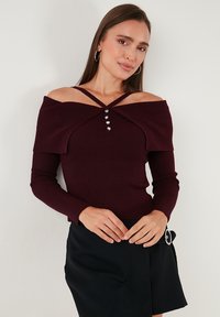 Sieviete valkā tumši violetu plecu izgriezumu svītrainu džemperi ar sirds formas pogām un melnu svārku ar sānu sprādzi.