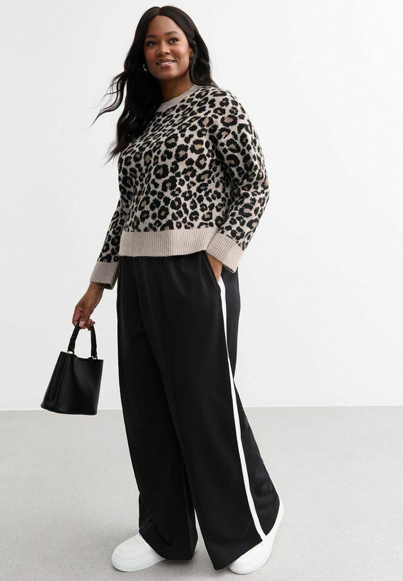 Un pull en imprimé léopard avec un col rond côtelé, associé à un pantalon large noir avec des rayures blanches sur les côtés et des baskets blanches. Petit sac à main noir.