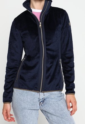 Personne portant une veste polaire bleu marine à fermeture éclair avec des poches zippées et un jean bleu clair, debout devant un fond uni.