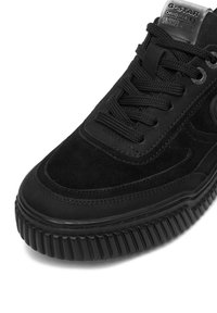 Čierna semišová teniska s textúrovanou gumovou podrážkou, okrúhlym špičkou a šnurovaným dizajnom. Má logo na jazyku a švy ako detaily.