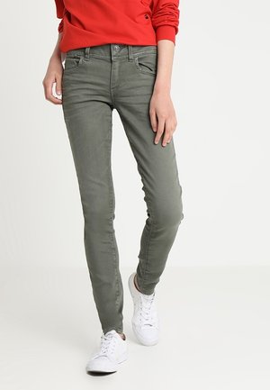 Jeans skinny verde oliva con una textura suave, diseño de cinco bolsillos y costuras en contraste, combinados con zapatillas blancas.