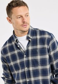 Homme aux cheveux bruns courts portant une chemise à carreaux bleus et blancs boutonnée par-dessus un t-shirt blanc à col rond, regardant vers sa gauche.