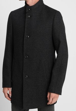 Black textured men's coat with a high collar and five black buttons, worn over dark pants, shown from neck to mid-thigh.