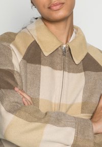 Villasekoitetehty takki, jossa on suuri kaulus ja ruudullinen kuvio beigeä, ruskeaa ja kermaa. Takissa on vetoketjusuljenta ja teksturoitu pinta.