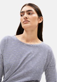 Frau mit langen braunen Haaren, die ein weiß-blau gestreiftes Shirt trägt und nach links schaut, vor einem schlichten weißen Hintergrund.