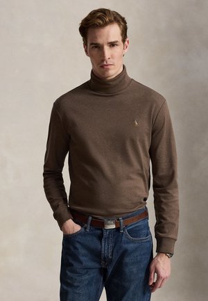 Jeune homme portant un pull à col roulé marron avec un logo, un jean bleu et une ceinture en cuir marron, debout devant un fond uni.
