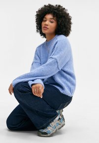 Hellblaues Strickpullover mit langen Ärmeln, kombiniert mit dunklen Jeans und grauen Sportschuhen mit türkisfarbenen Akzenten.