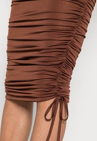 Těsná hnědá sukni s řasením a bočním stahováním do mašle, nošená nad kolena, odhalující část holých nohou na jednobarevném pozadí.