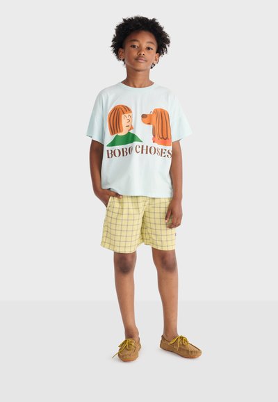 Chłopiec z kręconymi włosami, ubrany w jasnoniebieską koszulkę z kreskówką przedstawiającą dziewczynkę i psa oraz żółte, kratkowane spodenki, stoi z ręką w kieszeni.