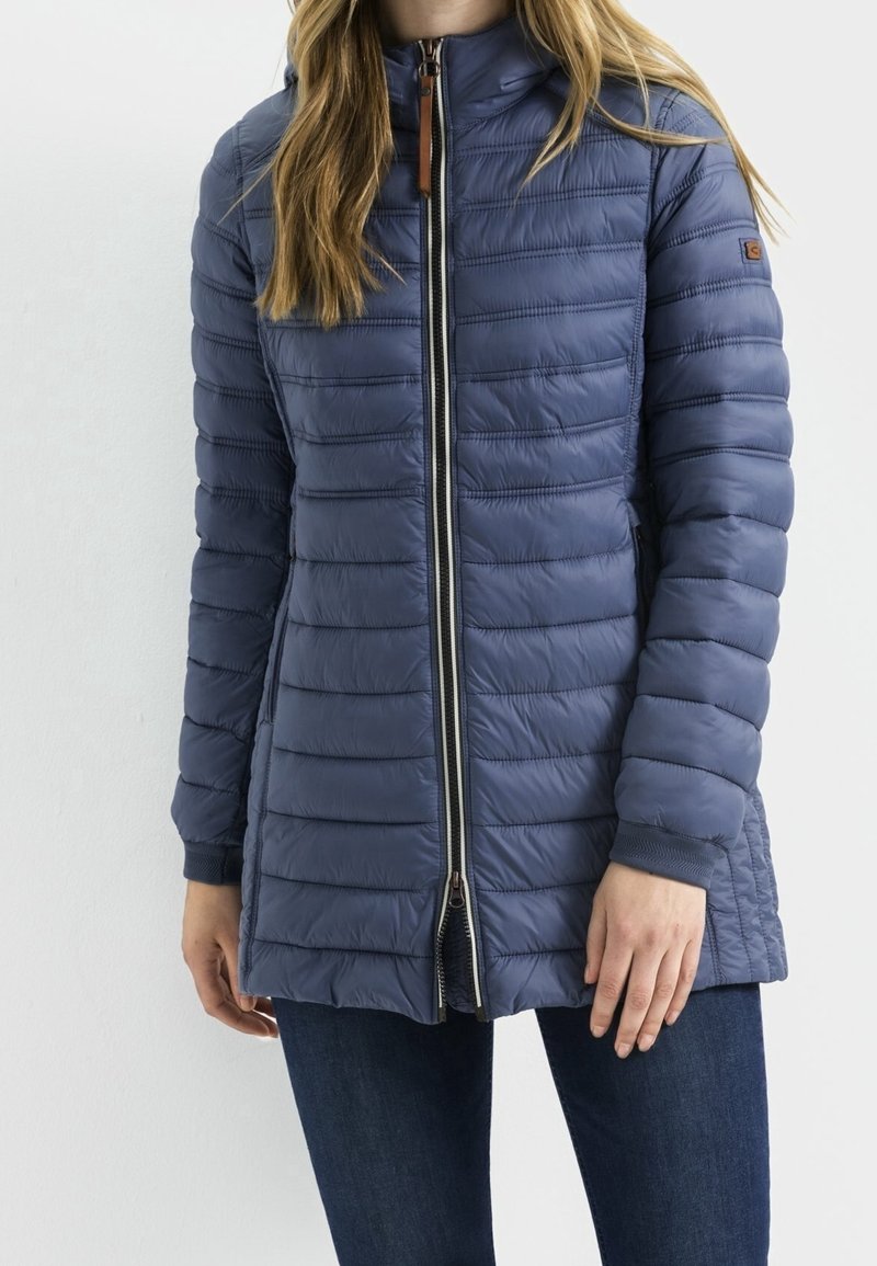 Femme portant une veste matelassée bleu marine mi-longue avec une fermeture éclair frontale et des poches latérales, associée à un jean bleu foncé.