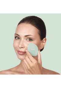 Outil de massage facial en silicone de couleur vert pâle, en forme ovale, utilisé sur la peau. Le nom de la marque "GESKE" est visible en texte blanc.