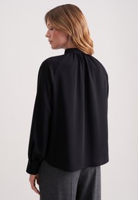 Blusa nera con collo alto, vestibilità rilassata e maniche lunghe. Realizzata in tessuto morbido con pieghe sulla schiena e polsini bottone.