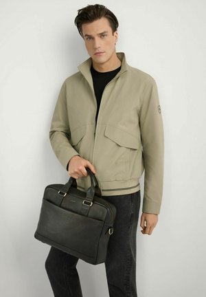 Młody mężczyzna w beżowej kurtce i czarnej koszuli, trzymający czarną skórzaną teczkę na jednolitym białym tle.