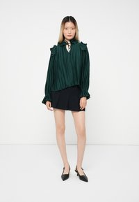 Blusa verde plissettata con spalle a ruota e scollo a V. Abbinata a una minigonna nera plissettata e scarpe nere a punta.