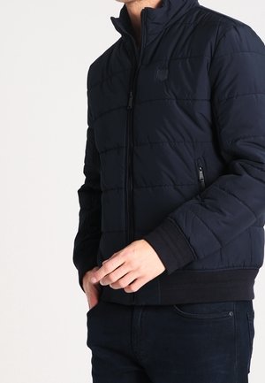 Mann trägt eine marineblaue gesteppte Jacke mit Reißverschluss vorne und Reißverschlusstaschen an den Seiten, steht mit einer Hand nahe der Taille.