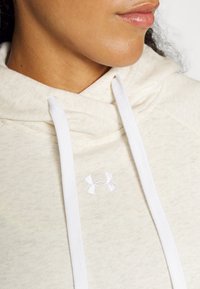 Felpa crema con cordini bianchi, con un piccolo logo bianco sul davanti. Il tessuto appare morbido con una texture sottile.