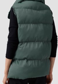 Grüne gepolsterte Weste mit hohem Kragen, gestepptem Design und glatter Textur. Der Rücken ist etwas länger und hat einen abgerundeten Saum, ohne sichtbare Beschläge.