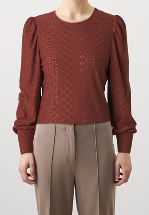 Kobieta ubrana w rdzawe brązową, koronkową bluzkę z długim rękawem, z bufkami na ramionach oraz beżowe spodnie z wysokim stanem na jednolitym tle.
