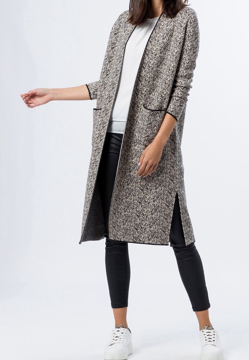 Długi, otwarty płaszcz z fakturowanym czarno-białym wzorem, z bocznymi rozcięciami i wąskimi kieszeniami, zestawiony z czarnymi legginsami i białymi trampkami.