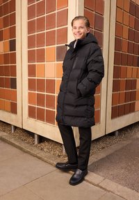 Doudoune noire avec capuche et poches latérales, portée sur un pantalon noir et des chaussures foncées. Debout devant un mur en briques à motif.