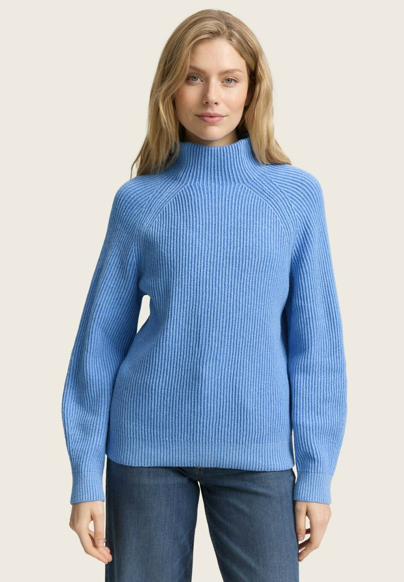 Pull en maille côtelée bleu clair avec un col montant, des épaules tombantes et des manches longues, associé à un jean foncé. Texture douce et coupe décontractée.