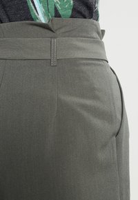 Šedé kalhoty s hladkou texturou, strukturovaným pasem a bočními švy kapes, doplněné vzorovaným černým topem.