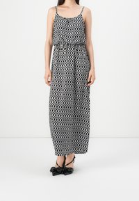 Robe maxi à motifs noir et blanc avec fines bretelles, taille froncée et silhouette fluide. Associée à des chaussures noires brillantes nouées en nœud.
