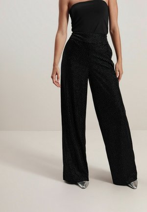 Femme portant un pantalon noir à jambes larges avec petits points blancs, un top noir sans bretelles, et des chaussures argentées pointues, debout devant un fond uni.