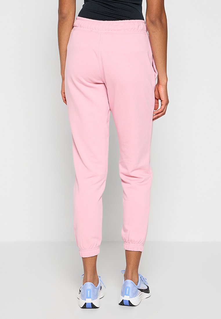 Kobieta nosząca jasnoróżowe dresy z elastycznymi mankietami i pasem, w połączeniu z jasnoniebieskimi i białymi butami do biegania, stojąca na jednolitym tle.