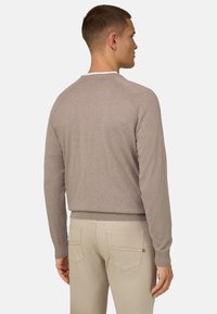 Homme en pull beige et pantalon beige clair vu de dos sur un fond blanc uni.