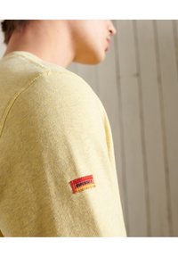 Felpa gialla in cotone con scollo rotondo e una piccola etichetta rossa e arancione cucita sulla manica con la scritta "Superdry".