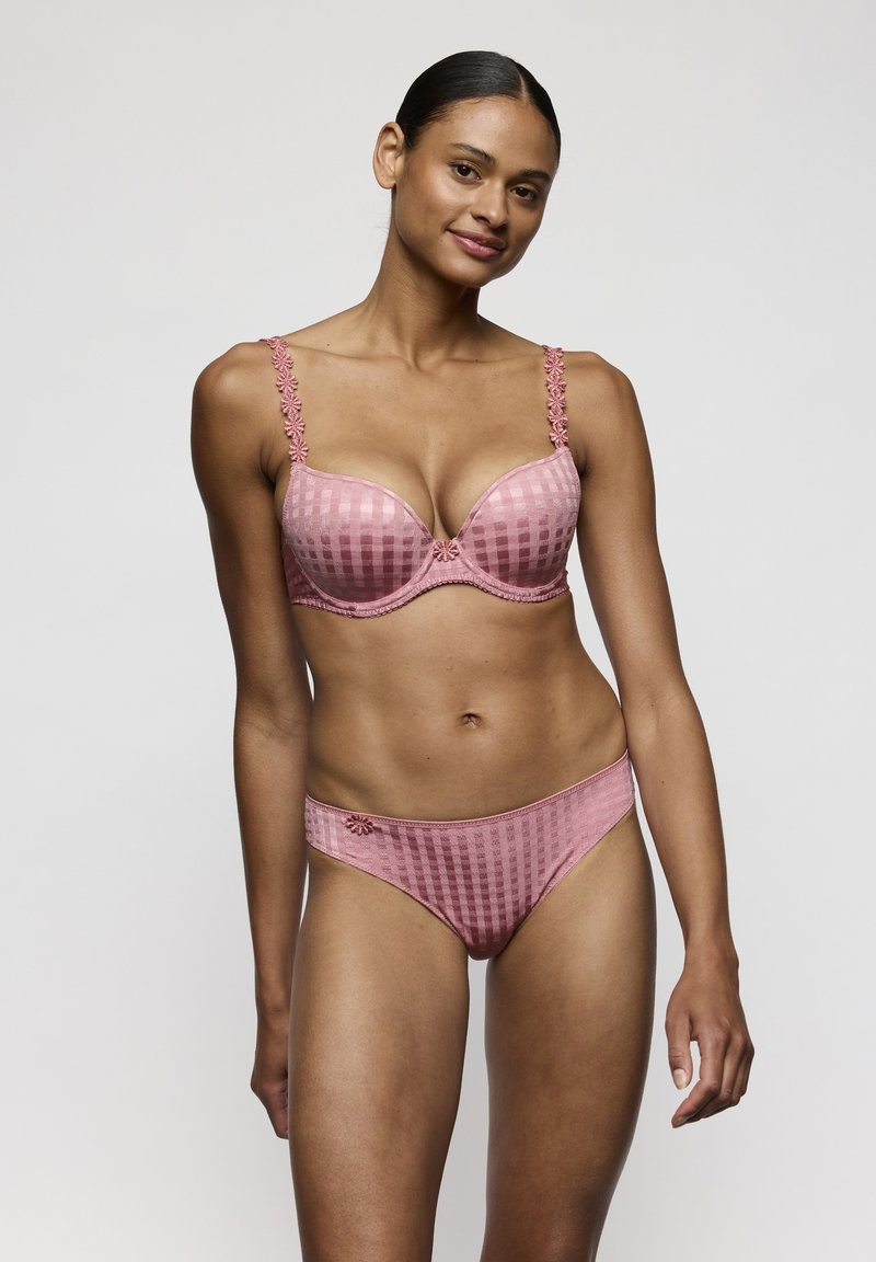 Femme debout portant un soutien-gorge et une culotte assortis à carreaux roses avec des détails de bretelles florales, sur un fond gris uni.