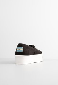 Chaussure en toile noire slip-on avec une semelle blanche épaisse et un petit logo TOMS sur le talon, vue de dos sur une surface blanche.