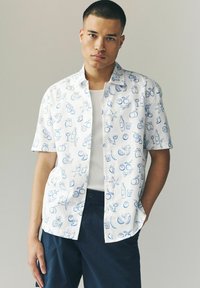 Chemise à manches courtes en tissu blanc avec des motifs dessinés à la main en bleu représentant des fruits et des boissons, associée à un t-shirt blanc uni et un pantalon sombre.