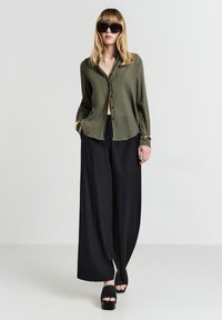 Donna con camicia button-down verde scuro, pantaloni neri a gamba larga, sandali con piattaforma neri, braccialetti dorati e grandi occhiali da sole neri su sfondo bianco.