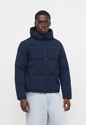 Uomo che indossa una giacca imbottita blu scuro con cappuccio e cerniera, in piedi su uno sfondo uniforme chiaro con jeans chiari.