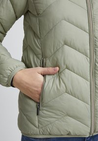 Veste matelassée vert clair avec fermeture éclair et poches latérales. Présente un motif en chevrons et des poignets élastiques pour un ajustement confortable.