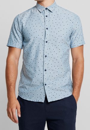Maglietta azzurra chiara a maniche corte con un motivo geometrico di piccoli triangoli neri e bianchi, dotata di colletto a camicia e bottoni scuri.