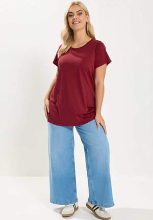 Mujer rubia con una camiseta burdeos holgada, vaqueros de pierna ancha azul claro y zapatillas blancas con rayas negras, de pie contra un fondo blanco.