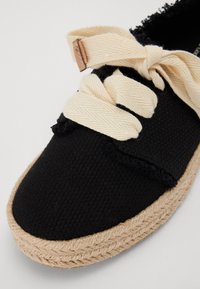 Espadrilla nera con texture intrecciata, lacci in tessuto crema e suola in juta intrecciata. Presenta un piccolo etichetta in pelle al punto di connessione dei lacci.