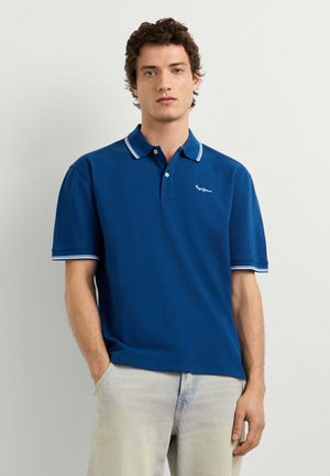 Junger Mann steht mit den Händen in den Taschen, trägt ein blaues Polohemd mit weißem Rand und helle Jeans vor einem einfarbigen Hintergrund.