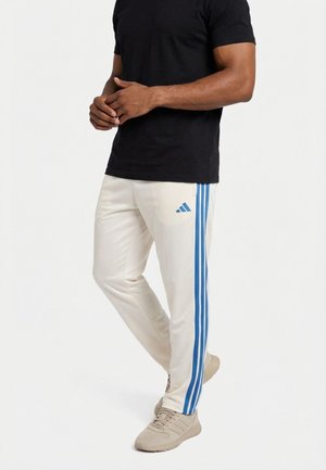 Hombre de pie con las manos entrelazadas, vistiendo pantalones deportivos blancos con franjas azules y una camiseta negra, zapatillas beige sobre un fondo blanco.