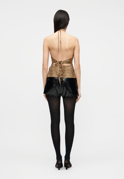 Femme aux longs cheveux foncés portant un haut dos nu à imprimé léopard, un short en cuir verni noir, des collants noirs et des talons noirs, debout.